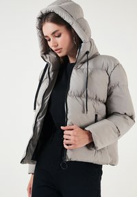 Svetlošedá puffová bunda s kapucňou, s prešívaným dizajnom, predným zipsom a nastaviteľnými šnúrkami. Jednoduchý a funkčný štýl.