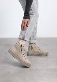 Zapatillas altas beige de material de ante texturizado, con cierre de cordones en la parte delantera, cremallera lateral y suela gruesa y estriada. Diseño casual con jeans arremangados.