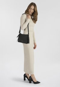 Schwarze Leder-Schultertasche mit verstellbarem Riemen, rechteckiger Form, strukturiertem Oberflächen und seitlichen Akzentquasten, kombiniert mit einem leichten Kleid.
