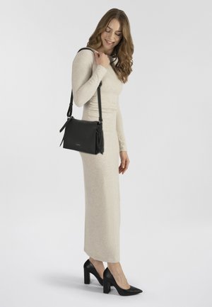 Schwarze Leder-Schultertasche mit verstellbarem Riemen, rechteckiger Form, strukturiertem Oberflächen und seitlichen Akzentquasten, kombiniert mit einem leichten Kleid.