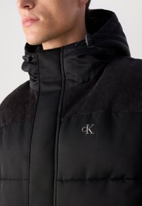 Jacke acolchoada preta com um colarinho alto, ombros de corduroy texturizado e um pequeno logo em relevo no peito. Cordões no capuz.