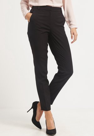 Femme portant un pantalon noir slim, un chemisier rose clair et des chaussures à talons noires, debout avec une main dans la poche.