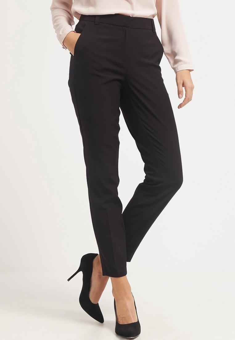 Femme portant un pantalon noir slim, un chemisier rose clair et des chaussures à talons noires, debout avec une main dans la poche.