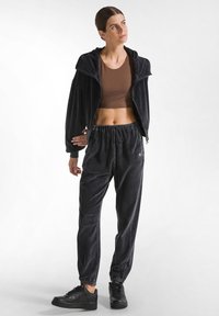 Schwarzes Velours-Trainingsanzug mit einer Reißverschluss-Kapuzenjacket und elastischen Hosen. Ein braches Crop-Top und schwarze Turnschuhe runden den Look ab.