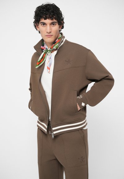 Jovem com cabelo encaracolado, vestindo uma jaqueta castanha com fecho e calças combinando, uma camisa branca e um cachecol colorido, de pé com as mãos nos bolsos.