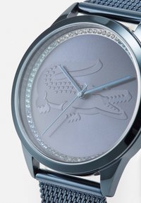 Montre-bracelet en acier inoxydable avec un cadran gris lisse, un anneau en cristal, un logo en relief et un bracelet en métal maillé. Dispose de aiguilles minimalistes.