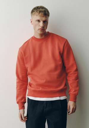 Junger Mann mit kurzen blonden Haaren trägt roten Sweatshirt über weißem Hemd und dunklen Hosen, steht vor einfarbiger heller Wand.