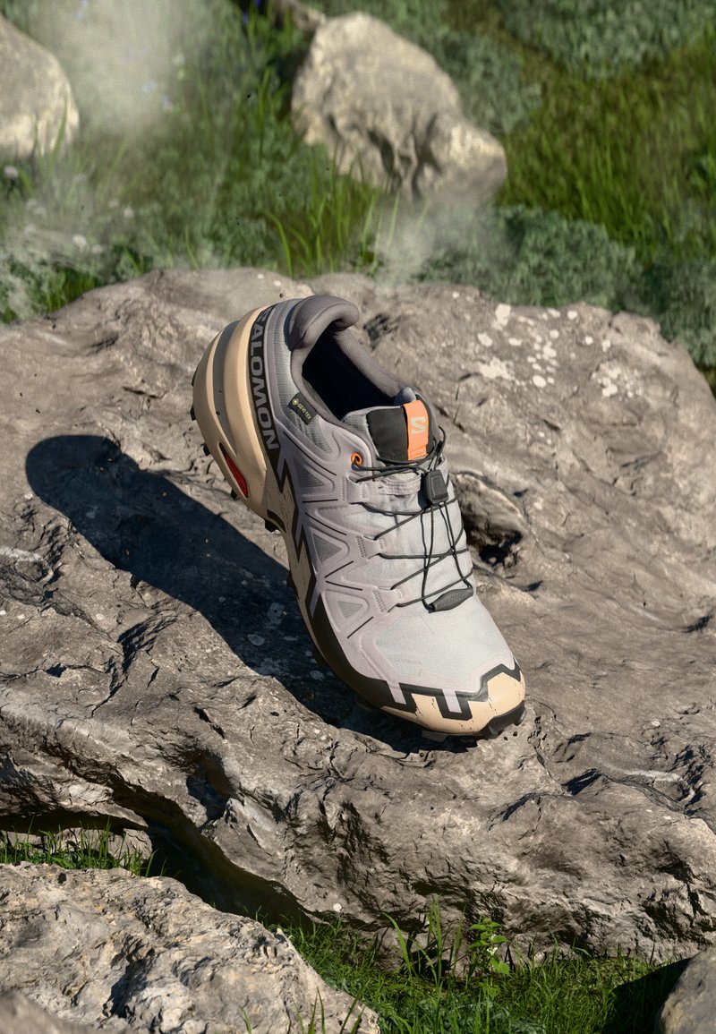 Lys grå terrengløpesko med beige detaljer, teksturert overdel, sorte snørebånd og markert mønster på sålen. Inkluderer merkevarelogo.