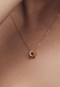 Złoty naszyjnik z misternie splątanym wisiorkiem, zakończony delikatnym łańcuszkiem. Gładka, błyszcząca faktura podkreśla elegancki design.