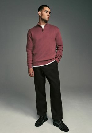 Marsala Zip-Kragenpullover, lange Ärmel, strukturiertes Gewebe. Kombiniert mit lockeren schwarzen Hosen und schwarzen klobigen Schuhen. Neutraler Hintergrund.
