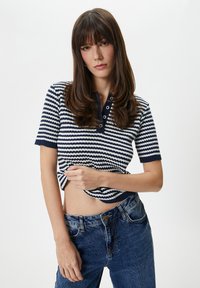 Stripet strikpullover i marineblå og hvid, kortærmet, med marineblå krave og tre-knap lukning. Båret med blå højtaljede jeans.