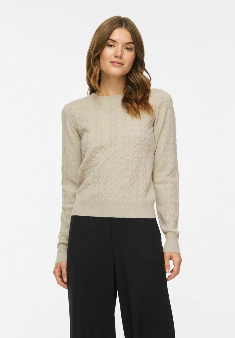 Femme aux cheveux châtain moyen portant un pull beige torsadé et un pantalon noir à jambes larges, debout devant un fond uni.