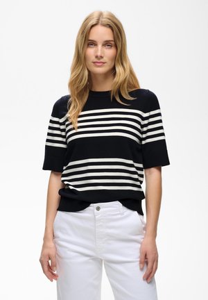 Blond vrouw met losse golven die een zwarte trui met korte mouwen en witte horizontale strepen draagt en witte broek, staand tegen een witte achtergrond.