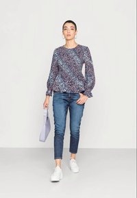Blusa peplum de manga longa com um padrão floral em azul-marinho, rosa e branco, combinada com jeans azuis e tênis brancos. Segura uma bolsa lilás clara.