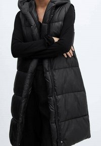 Veste sans manches noire matelassée avec capuche, dotée d'un design matelassé, fermeture éclair à l'avant et poches latérales. Le matériau est lisse et brillant.