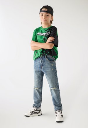 Chłopiec ubrany w zielono-czarną koszulkę Minecraft, podarte niebieskie jeansy, białe trampki i czarną czapkę z daszkiem na odwrót, stoi z założonymi rękami.