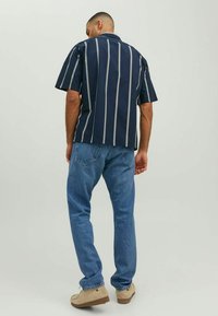 Tamsiai mėlyna marškiniai trumpomis rankovėmis su baltomis vertikaliomis juostelėmis, derinami su šviesiai mėlynomis džinsomis ir smėlio spalvos bateliais. Marškiniai turi klasiką apykaklę ir laisvą siluetą.