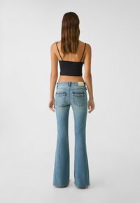 Jean flare bleu clair avec taille mi-haute, texture délavée et deux poches arrière. Porté avec un haut noir ajusté sur un fond neutre.