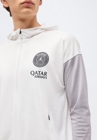 Νέος που φοράει λευκό και γκρι φούτερ με φερμουάρ, με το λογότυπο της Παρί Σεν Ζερμέν και τον χορηγό Qatar Airways στο στήθος.