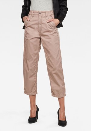 Kvinde iført cropped beige bukser med elastiske manchetter, sorte høje hæle, grå top og sort jakke, hænderne i lommerne.