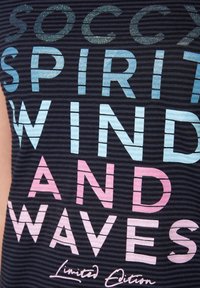 Modrý a růžový glitrový text na tmavé pruhované tkanině. Text zní "SOCCY SPIRIT WIND AND WAVES" s detailem "Limitovaná edice".