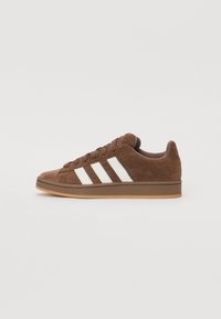 Brown suede sneaker with white three-stripe design, flat rubber sole, and rounded toe. Features matching laces and minimal branding.