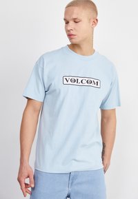 Camiseta de algodón azul claro con mangas cortas, que presenta un logo rectangular negro con la palabra "VOLCOM" en letras negritas en el frente.