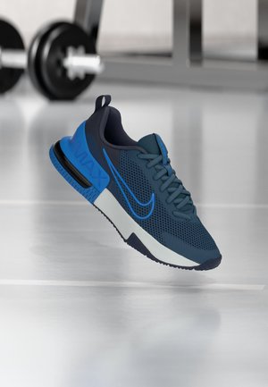 Chaussure de sport bleue et noire avec dessus en mesh et semelle à air visible, flottant au-dessus du sol de la salle de sport avec une barre en arrière-plan.
