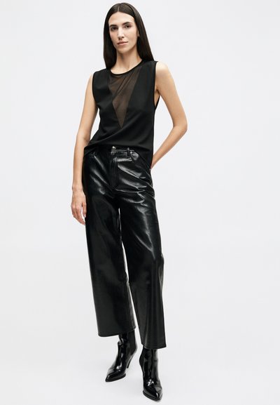 Woman with long dark hair wearing sleeveless black top with sheer V detail, black leather wide-leg pants, and black pointed boots.