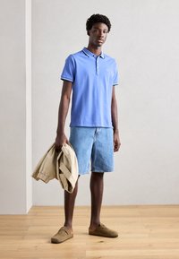 Svetlomodrá polo košeľa s golierom a gombíkmi, skombinovaná s džínsovými šortkami. V ruke drží béžovú bundu, na nohách má hnedé slip-on topánky.