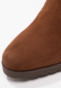 Bottine en daim marron avec un bout arrondi et une surface texturée. La semelle est foncée et en caoutchouc. Présente une finition lisse et un design robuste.