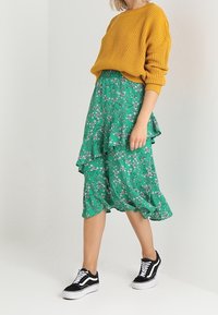 Woman wearing mustard yellow knit sweater, green floral midi skirt with ruffles, and black-white sneakers walking against white background.