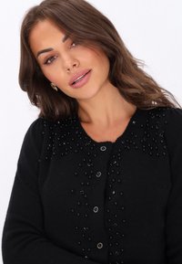 Juodas kardiganas su tekstūruota paviršiumi, puošniomis juodomis karoliukais ir dekoratyviais mygtukais. Apvalus iškirpimas; prigludęs dizainas. Minkšta medžiaga.