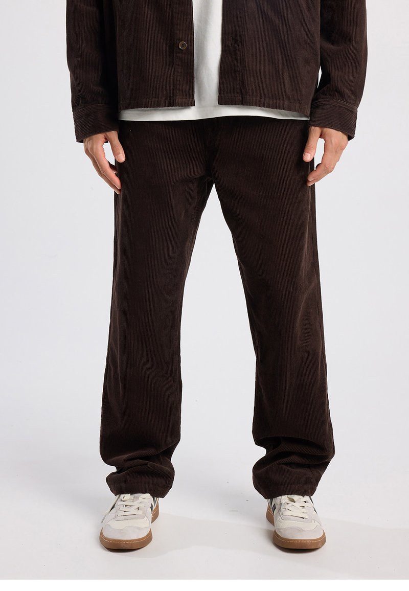 Bruin corduroy jack en broek set met geribbelde textuur; ontspannen pasvorm; gecombineerd met een wit T-shirt en lichtgekleurde sneakers.