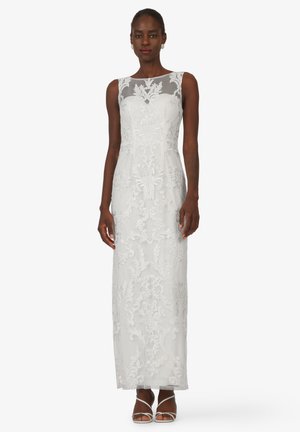 Weißes bodenlanges Kleid mit floralem Stickerei, einem transparenten Ausschnitt und einer taillierten Silhouette. Das Model trägt offene, weiße High Heels.