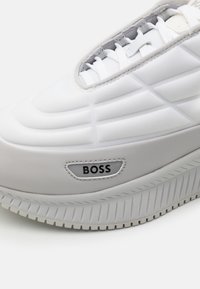 Bílá teniska s hladkou texturou, strukturovaným designem, výraznou podrážkou s vroubkovaným vzorem a logem "BOSS" na boku.