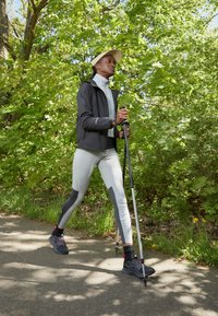 Mujer caminando por un sendero con bastones de trekking, vistiendo una chaqueta deportiva, leggings, zapatillas y visera en un entorno al aire libre verde y frondoso.