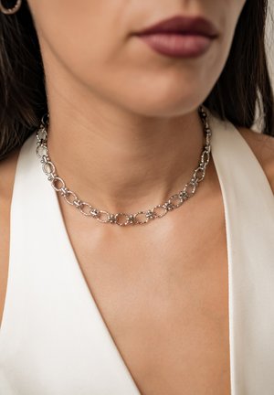 Silberne Kette mit ovalen Gliedern, getragen am Hals einer Person mit weißem ärmellosem Oberteil.