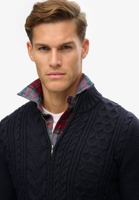 Pullover blu marina a maglia con zip sul colletto, indossato su una camicia a quadri rossa e blu, con un colletto girocollo bianco sotto.