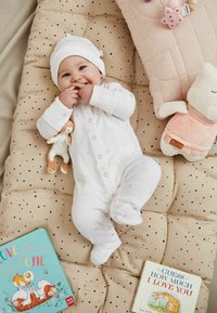 Witte romper met roze patronen, een witte muts en een pluche speelgoed, liggend op een beige kussenoppervlak met zwarte stippen, omringd door kinderboeken.