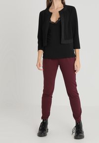 Černá zkrácená bunda s texturovanými lemy, přes černou krajkovou košilku, spárovaná s úzkými burgundskými kalhotami a černými šněrovacími botami.