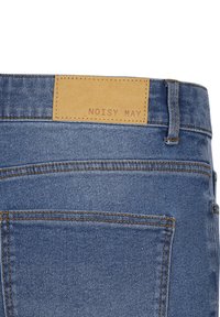 Lichte spijkerbroek van stevig materiaal, zichtbaar stiksel en een bruine leren label met "NOISY MAY" in reliëf.