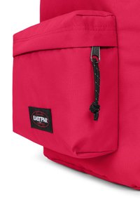 Röd tygryggsäck med en framficka med dragkedja, svart hårdvara och en märkesetikett med en jordglobdesign. Tillverkad av hållbart material.