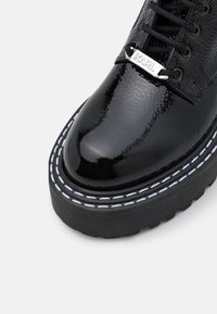 Black patent leather ankle boot with a thick black sole, white stitching detail, and a silver accent labeled "KARL." Laced design with eyelets.
