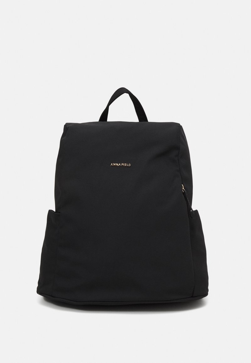 Sac à dos noir minimaliste avec poignée supérieure, poches latérales et petit logo doré "ANNA FIELD" sur le devant.