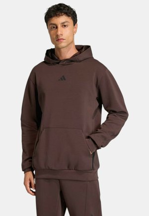 Giovane uomo che indossa una felpa con cappuccio Adidas marrone scuro e pantaloni abbinati, in piedi con una mano in tasca su uno sfondo semplice.