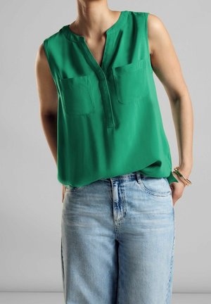 Grøn ærmeløs bluse med V-udskæring og to brystlommer, parret med lyseblå denimjeans, der viser en afslappet pasform og glat tekstur.