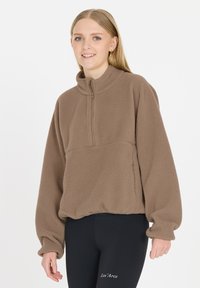 Brauner Fleece-Pullover mit hohem Kragen und Viertel-Reißverschluss, ausgestattet mit einer strukturierten Oberfläche und lockerer Passform, kombiniert mit schwarzen Leggings.