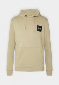 Sudadera beige hecha de tejido suave, con un bolsillo frontal, capucha ajustable con cordón y un parche cuadrado negro con texto en el pecho.