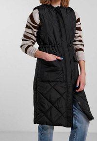 Svart vadderad ärmlös kappa med dragsko i midjan, som har två sidofickor och en fullständig dragkedja fram, bärs över en randig tröja.